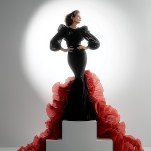 May include: A black gown with voluminous sleeves and a fitted silhouette. The gown features a dramatic red ruffled train. The model is standing on a white platform against a bright, white background.