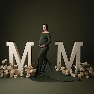 May include: A pregnant person in a dark green off-the-shoulder gown poses in front of large white letters spelling "MM". The letters are surrounded by cream-colored roses and greenery, set against a dark green backdrop.
