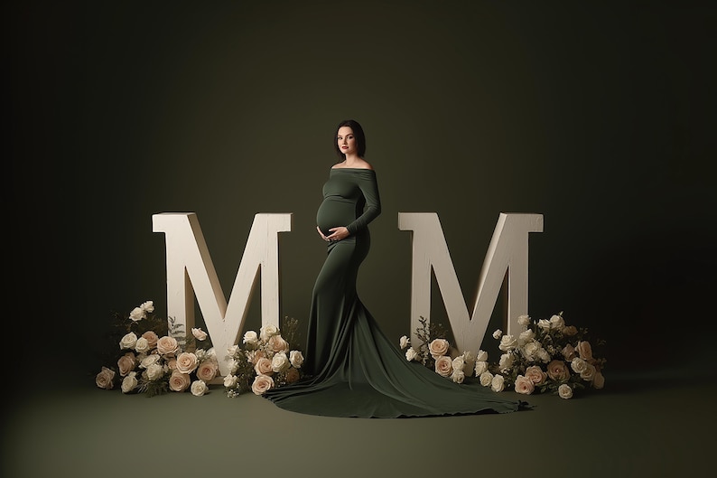 May include: A pregnant person in a dark green off-the-shoulder gown poses in front of large white letters spelling "MM". The letters are surrounded by cream-colored roses and greenery, set against a dark green backdrop.