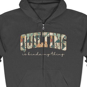 May include: Dark gray zip-up hoodie with the word "QUILTING" in a patchwork design across the chest. Below the word is the phrase "is kinda my thing" in a script font. The hoodie has a drawstring hood and two front pockets.