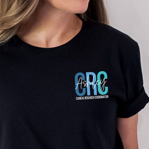 May include: A black short-sleeved t-shirt featuring a graphic on the left chest. The design includes the letters CRC in blue and teal, with the name Ashley written in white script across them and the text CLINICAL RESEARCH COORDINATOR below.