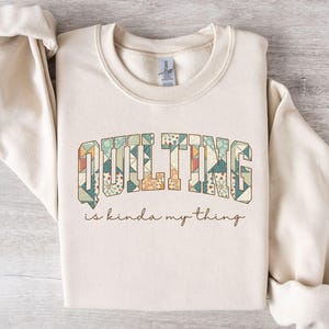 May include: Sand-colored sweatshirt with the text "QUILTING" in a patchwork design, and the phrase "is kinda my thing" in a script font. The sweatshirt has a crew neck and long sleeves.