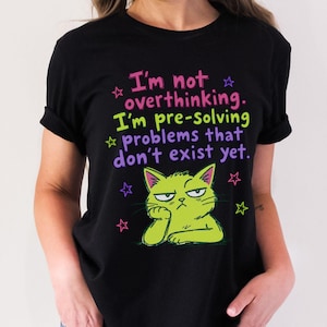 May include: A black short-sleeved t-shirt featuring a graphic of a lime green cat with a grumpy expression. The text reads: I'm not overthinking. I'm pre-solving problems that don't exist yet. The design includes small pink and green stars.