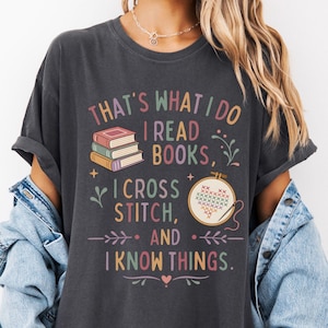 May include: Dark gray t-shirt with the text "THAT'S WHAT I DO I READ BOOKS, I CROSS STITCH, AND I KNOW THINGS." The shirt features illustrations of books and cross-stitch embroidery. The shirt is a comfortable, casual style.