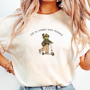 May include: An ivory short-sleeved t-shirt featuring a graphic of a green frog wearing sunglasses and a cap while riding a scooter. The text above the frog reads, Off to commit aunt foolery. The shirt has a crew neckline and a relaxed fit.