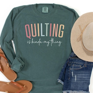 May include: A blue spruce long-sleeve shirt with the text "QUILTING is kinda my thing" in colorful letters. The shirt is paired with denim shorts, a tan hat, and brown sandals. The image is styled on a white background.
