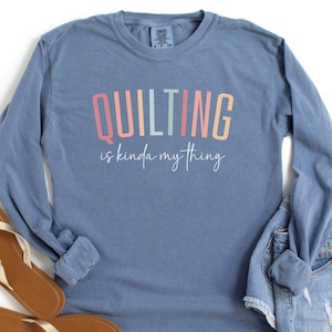 May include: A long-sleeved, blue shirt with the text "QUILTING is kinda my thing" in colorful letters. The shirt is paired with denim shorts and brown sandals with gold straps. The words "blue jean" are written in the bottom left corner.