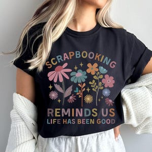 May include: A black short-sleeve t-shirt featuring a colorful floral graphic and the text "SCRAPBOOKING REMINDS US LIFE HAS BEEN GOOD" in a multi-colored font. The design includes various stylized flowers, leaves, and small star accents.