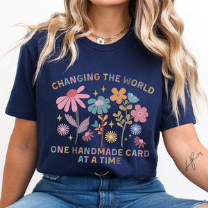 May include: A set of three ceramic mugs with a speckled, cream-colored glaze. Each mug features a different hand-painted design: a blue and green leaf, a yellow flower, and a red berry. The mugs have a simple, modern design.