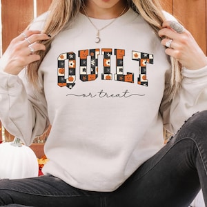 May include: A sand-colored crewneck sweatshirt featuring the text "QUILT or treat" in a blocky, patterned font. The letters are filled with a checkered design containing orange pumpkins, white flowers, and black accents. The text is printed in black.