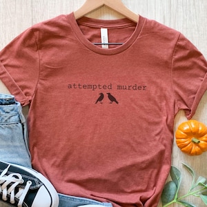 Pu&ograve; includere: T-shirt color ruggine con la scritta "attempted murder" e due illustrazioni di corvi. La maglietta &egrave; abbinata a jeans azzurri e scarpe da ginnastica Converse nere. Una piccola zucca e del verde completano l'ensemble a tema autunnale.