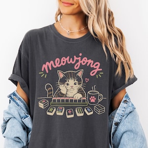 May include: Dark gray t-shirt with the text "meowjong" in pink script above a cartoon cat playing mahjong. The cat is brown and gray, with a paw print on the coffee mug. Several mahjong tiles are visible.