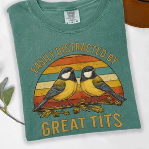 May include: Light green t-shirt with the text "EASILY DISTRACTED BY GREAT TITS" in a retro design. The graphic features two yellow and black birds perched on a branch against a sunset background. The shirt is a Comfort Colors brand.