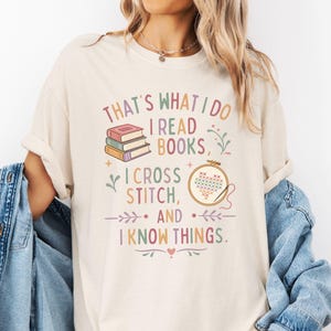 May include: Ivory-colored t-shirt with the text "THAT'S WHAT I DO I READ BOOKS, I CROSS STITCH, AND I KNOW THINGS." The design includes stacked books, a cross-stitch hoop, and decorative floral elements. The shirt is a relaxed fit.