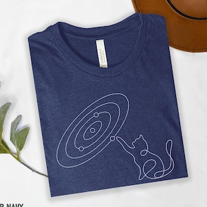 May include: Heather navy blue t-shirt with a white line drawing of a cat reaching towards a solar system design. The shirt is folded, with the text "HEATHER NAVY" visible at the bottom left.