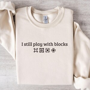 May include: A cream-colored sweatshirt with the text "I still play with blocks" in black, along with three quilt block designs. The sleeves are cuffed. The sweatshirt is on a light wood-grain surface.