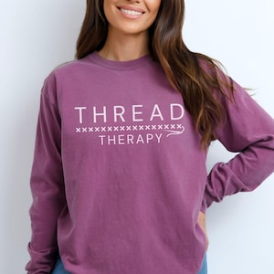 May include: A long-sleeved, purple t-shirt with the text "THREAD THERAPY" and a row of "x" symbols above the text.