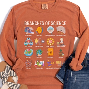 May include: An orange long-sleeve shirt with the text "BRANCHES OF SCIENCE" and illustrations of different scientific fields. The shirt is paired with denim shorts and sandals.