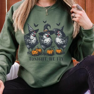 May include: Military green sweatshirt featuring a graphic of three chickens wearing witch hats, with pumpkins and bats. The text "TONIGHT, WE FLY" is printed below the chickens. The sweatshirt is a solid color.