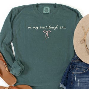 May include: A teal long-sleeve shirt with the text "in my sourdough era" and a pink bow graphic. The shirt is paired with brown sandals, denim shorts, and a beige hat. The words "blue spruce" are at the bottom left.