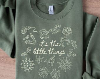 May include: Military green sweatshirt with the phrase "it's the little things" and various microbe illustrations in white. The long-sleeved crewneck top is folded on a wooden surface.
