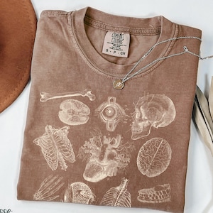 May include: Espresso-coloured t-shirt with a detailed anatomical print of bones, a heart, a brain, and other organs. The shirt has a crew neck and short sleeves. A silver necklace with a small pendant rests on the shirt. The shirt is labelled "Comfort Colors".