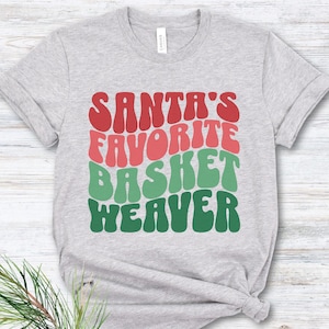May include: Athletic heather gray t-shirt with the text "SANTA'S FAVORITE BASKET WEAVER" in red, pink, and green. The shirt is a crew neck and has short sleeves. A sprig of pine and a pinecone are in the lower left corner.