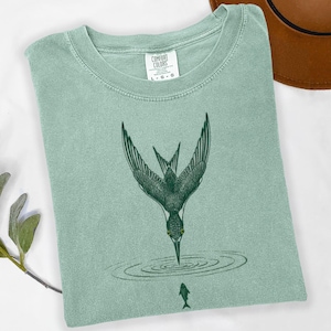 May include: A light green t-shirt with a crew neck featuring a dark green illustration of a bird diving into water to catch a fish. The shirt has a Comfort Colors tag.