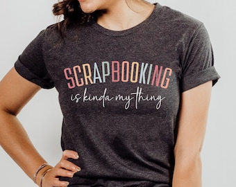 Può includere: Una maglietta color grigio scuro melange con il testo "Scrapbooking is kinda my thing" in colori arcobaleno.