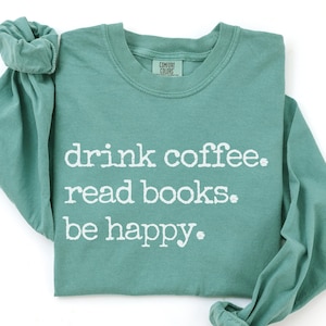 May include: A long-sleeve crewneck sweatshirt in a muted light green color. The front features white, distressed-style text that reads: drink coffee. read books. be happy. The garment has a Comfort Colors brand label at the inner neckline.