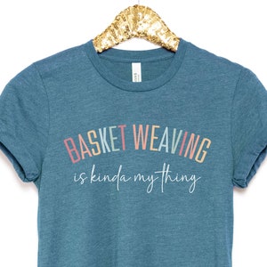 May include: A teal blue t-shirt with the text "Basket Weaving is kinda my thing" in a rainbow arch design.