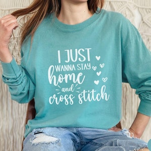 May include: A teal blue long-sleeved sweatshirt with the text "I just wanna stay home and cross stitch" in white lettering with small white hearts.