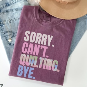 May include: A berry-colored t-shirt with the text "SORRY. CAN'T. QUILTING. BYE." in white, pink, and blue. The shirt is paired with light-wash denim jeans, a tan hat, and a gold necklace. The shirt size is XL 10-20.