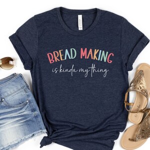 May include: A navy blue t-shirt with the text "Bread Making is kinda my thing" in rainbow colors. The shirt is on a white background with a pair of denim shorts and sandals.