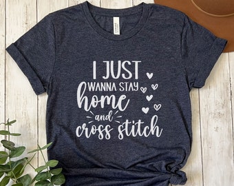 Op de afbeelding: Heather navy t-shirt met de witte tekst "I just wanna stay home and cross stitch". Het ontwerp bevat hartvormen. Het t-shirt heeft een ronde hals en korte mouwen.