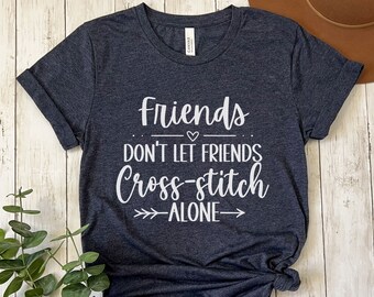 May include: A dark heather navy blue t-shirt with the text "Friends don't let friends cross-stitch alone" in white. The text is in a handwritten font with a heart symbol above the word "friends".