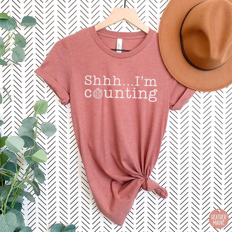 May include: A heather mauve t-shirt with a white graphic that says "Shhh... I'm counting" with a small clock icon.
