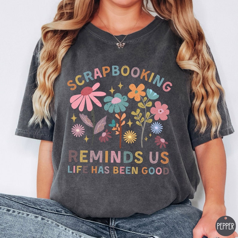 May include: A charcoal gray short-sleeve t-shirt featuring a colorful floral graphic and the text "SCRAPBOOKING REMINDS US LIFE HAS BEEN GOOD". The design includes various stylized flowers in shades of pink, blue, orange, and yellow, accented with small star shapes.