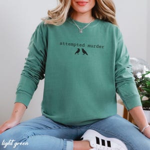 May include: Light green long-sleeve sweatshirt with the text "attempted murder" above two black bird silhouettes. The sweatshirt is a solid color and has a relaxed fit. The words "light green" are at the bottom left.