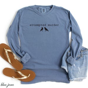 May include: A blue long-sleeve shirt with the text "attempted murder" and two black bird silhouettes. The shirt is paired with brown sandals and denim shorts.