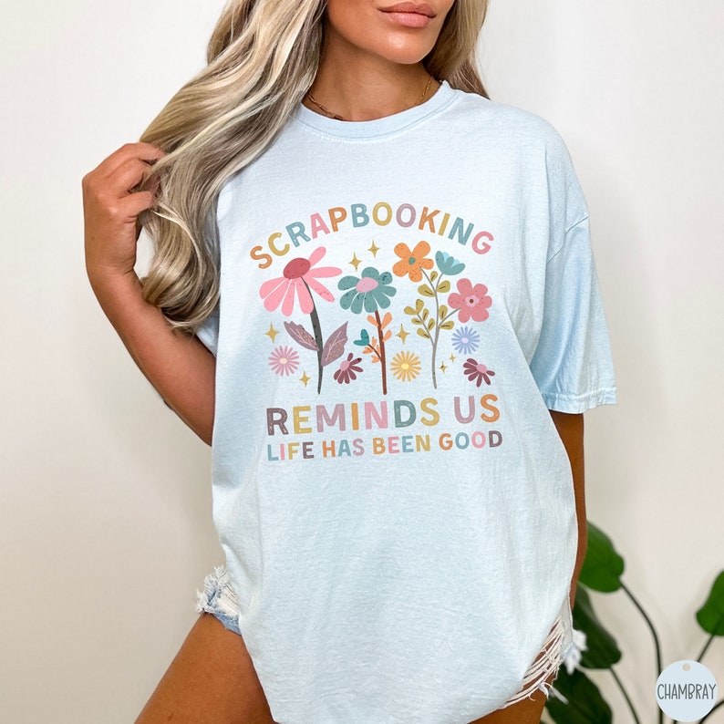 May include: A light blue t-shirt featuring a colorful floral graphic and the text "SCRAPBOOKING REMINDS US LIFE HAS BEEN GOOD" in a multi-colored, retro-inspired font. The design includes various stylized flowers, leaves, and small star accents.