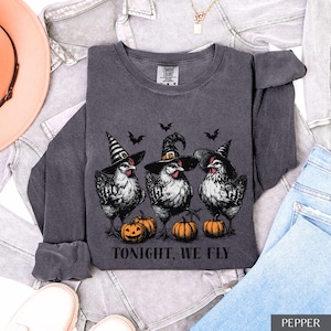 May include: A charcoal gray long-sleeve crewneck sweatshirt featuring a graphic of three chickens wearing black witch hats. The chickens stand above three orange pumpkins. The text "TONIGHT, WE FLY" is printed in black below the pumpkins.