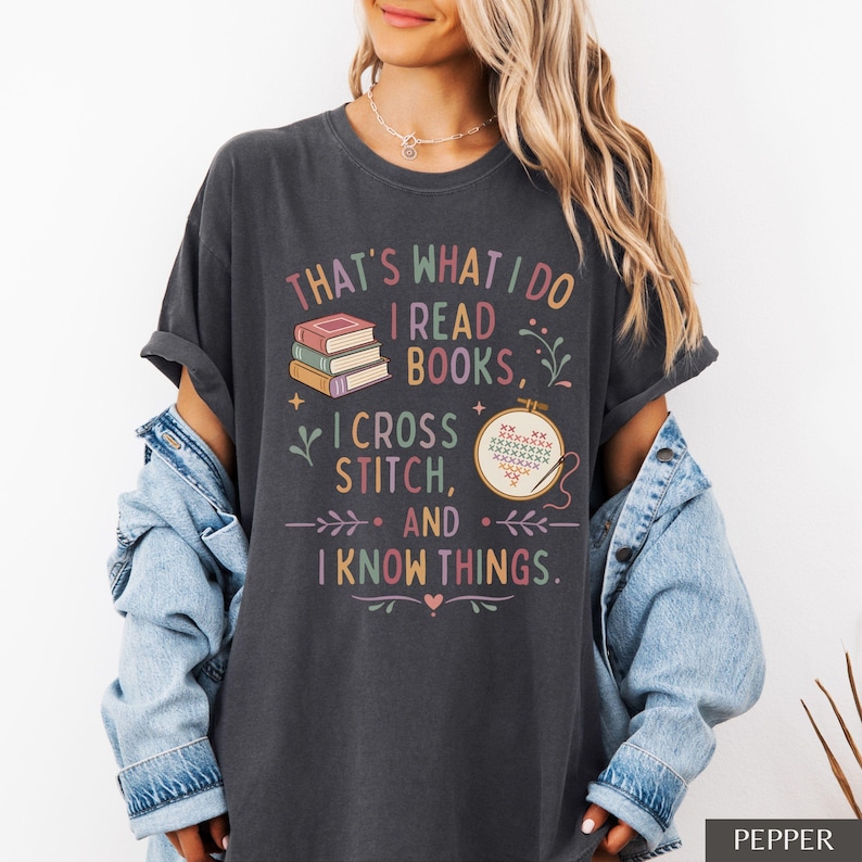 May include: Dark gray t-shirt with the text "THAT'S WHAT I DO I READ BOOKS, I CROSS STITCH, AND I KNOW THINGS." The shirt features illustrations of books and cross-stitch embroidery. The shirt is a comfortable, casual style.