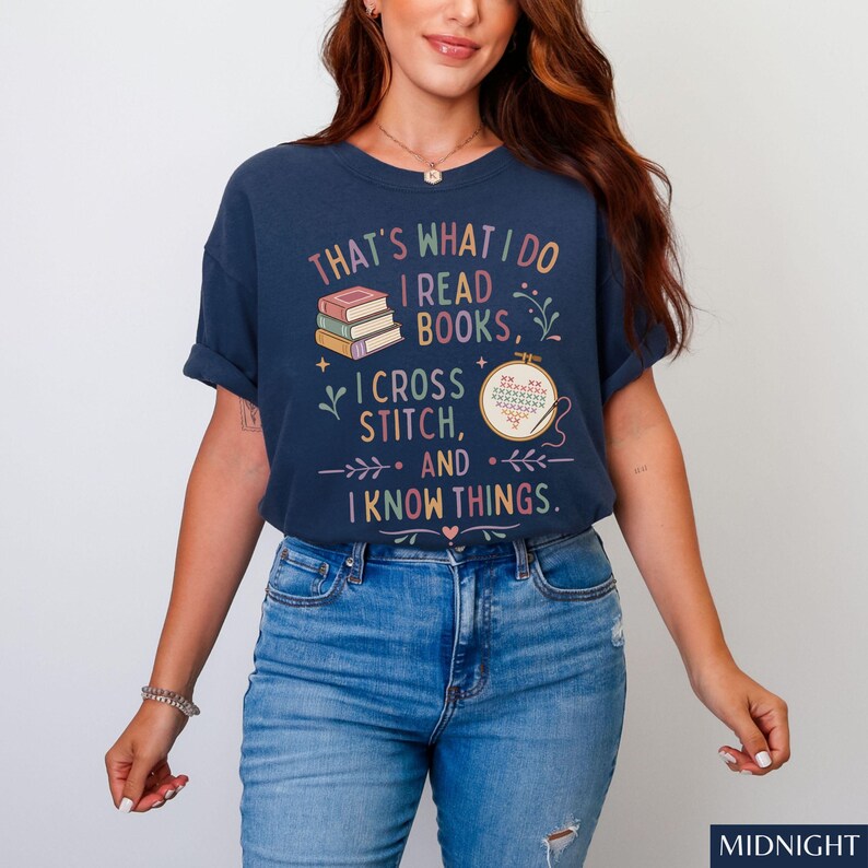 May include: Navy blue t-shirt with the text "THAT'S WHAT I DO I READ BOOKS, I CROSS STITCH, AND I KNOW THINGS." The graphic includes books and a cross-stitch design. The model is wearing blue jeans.