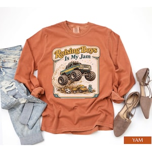 May include: A long-sleeved, rust-colored shirt featuring a graphic of a monster truck with the text "Raising Boys Is My Jam." The design includes a yellow and green monster truck, a soccer ball, and a small yellow truck.