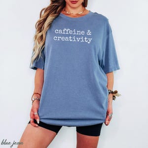 Pu&ograve; includere: Una maglietta blu jeans con le parole "caffeine & creativity" stampate in bianco. La maglietta &egrave; di vestibilit&agrave; ampia e ha maniche corte. La modella indossa pantaloncini neri.