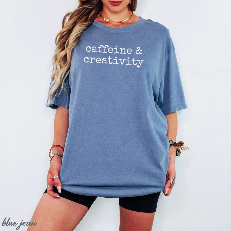 Pu&ograve; includere: Una maglietta blu jeans con le parole "caffeine & creativity" stampate in bianco. La maglietta &egrave; di vestibilit&agrave; ampia e ha maniche corte. La modella indossa pantaloncini neri.