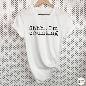 May include: White t-shirt with black text that reads "Shhh... I'm counting" and a small clock icon.