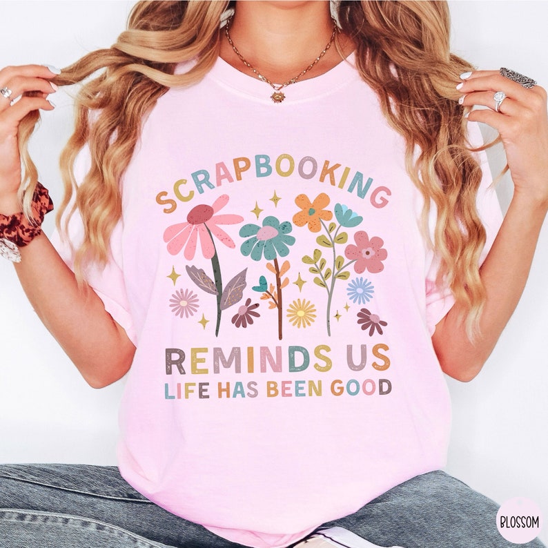May include: A light pink t-shirt featuring a colorful floral graphic and the text "SCRAPBOOKING REMINDS US LIFE HAS BEEN GOOD" in a multi-colored, retro-inspired font. The design includes various stylized flowers in shades of pink, blue, and orange.