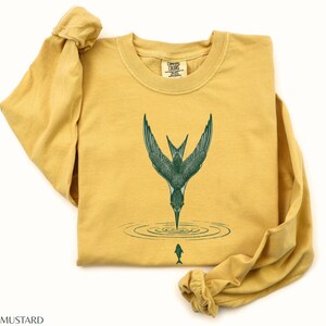 May include: Mustard-colored long-sleeve shirt with a green illustration of a bird diving into water to catch a fish. The shirt has a crew neck and the text "MUSTARD" at the bottom.